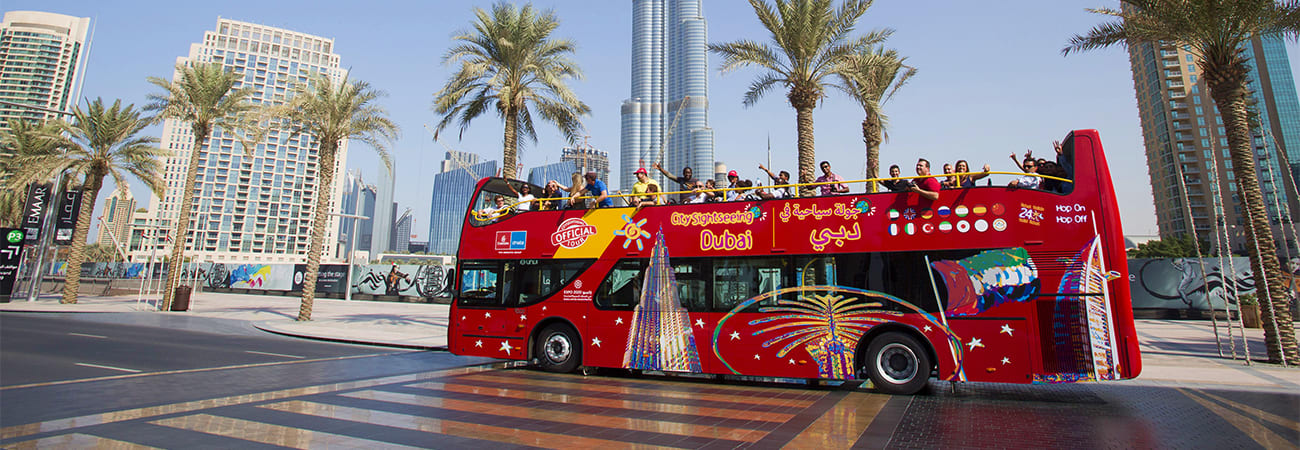 Red double-decker hop-on hop-off tour bus with passengers on upper deck, Burj Khalifa and palm trees in background, Dubai